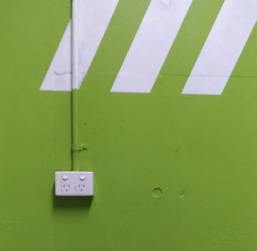 A bright green wall features a white electrical outlet with two sockets. Diagonal white stripes add a dynamic visual element above the outlet, and a vertical pipe runs down the wall, connecting to the outlet.