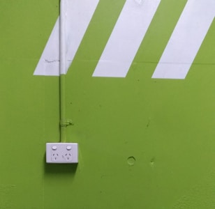A bright green wall features a white electrical outlet with two sockets. Diagonal white stripes add a dynamic visual element above the outlet, and a vertical pipe runs down the wall, connecting to the outlet.