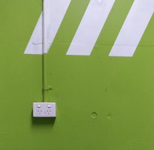 A bright green wall features a white electrical outlet with two sockets. Diagonal white stripes add a dynamic visual element above the outlet, and a vertical pipe runs down the wall, connecting to the outlet.