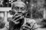 An elderly person is smoking a cigarette, holding it near their mouth with one hand, while the other hand holds a lighter. The person has a contemplative expression, with visible details in the face, and is dressed in a striped shirt. Smoke is softly rising around the face, and the background is blurred, suggesting an outdoor setting with trees.