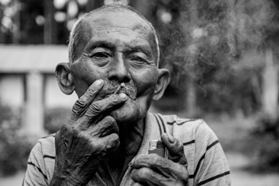 An elderly person is smoking a cigarette, holding it near their mouth with one hand, while the other hand holds a lighter. The person has a contemplative expression, with visible details in the face, and is dressed in a striped shirt. Smoke is softly rising around the face, and the background is blurred, suggesting an outdoor setting with trees.