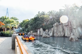 A bright, sunny scene featuring a river with yellow and black boats carrying passengers. The river is bordered by high rocky cliffs and lush green palm trees. In the distance, a modern bridge structure is visible, and the sun creates lens flares in the sky.