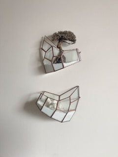 Two geometric glass terrariums are mounted on a light-colored wall. The upper terrarium contains a small bonsai tree planted in white sand. The lower terrarium has white stones or coral arranged in sand.