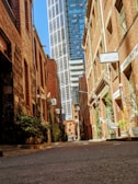 An urban alley lined with red brick buildings on both sides, leading towards a towering modern skyscraper in the background. The ground is paved, and various potted plants line the alleyway, adding a touch of greenery. There are signs along the buildings, indicating business names. The alley is bathed in bright sunlight, highlighting the textures of the brick walls.