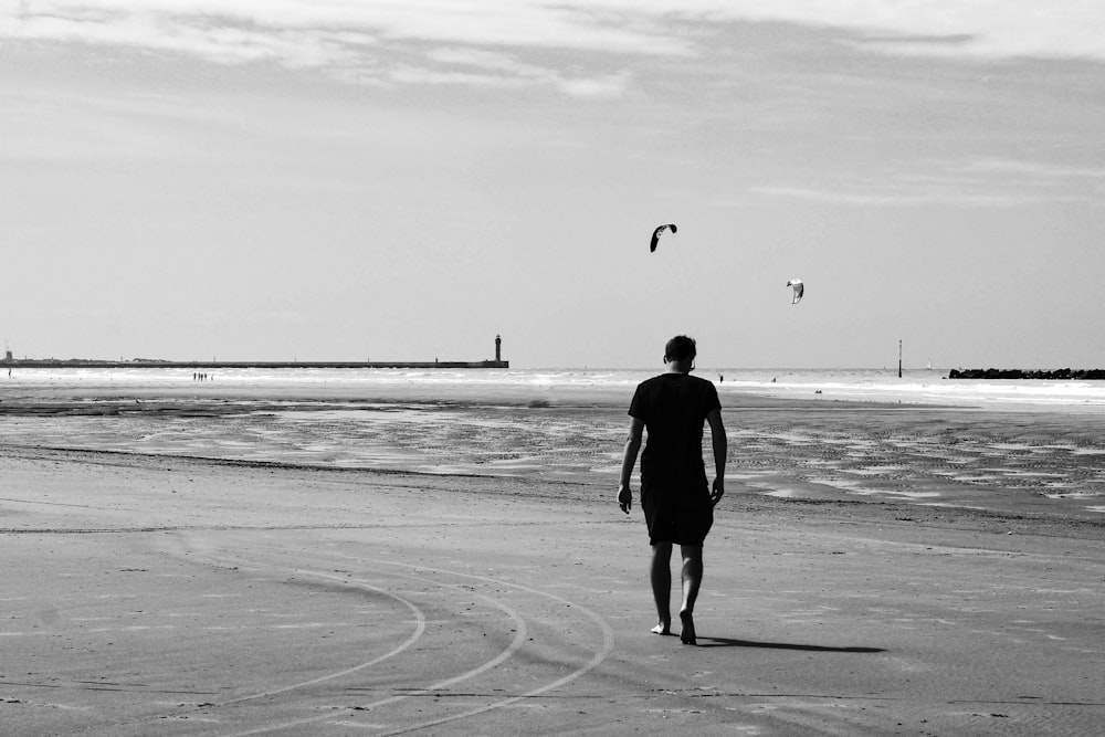 grayscale photo of man in black jacket walking on beach