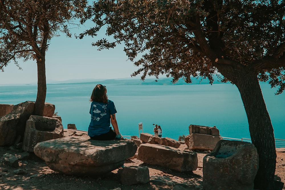 Frau im blauen Hemd sitzt tagsüber auf braunem Felsen in der Nähe von Wasser