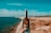 A hand holds a wine bottle labeled 'Talay' in the foreground near a rocky coastline. The background features several wind turbines in a row along the shore under a blue sky.