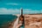 A hand holds a wine bottle labeled 'Talay' in the foreground near a rocky coastline. The background features several wind turbines in a row along the shore under a blue sky.