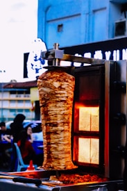 A vertical stack of seasoned meat heats on a rotating spit, surrounded by a glowing red-orange rotisserie grill. In the background, people sit at outdoor tables against a backdrop of urban buildings and signage.