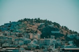 A hillside densely covered with colorful buildings with a large sign that reads 'KUŞADASI' on the right side. The top of the hill is lined with trees and a statue can be seen. The sky is clear, adding a serene backdrop to the lively urban area.