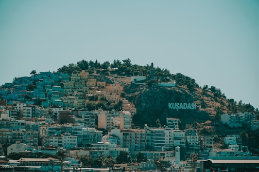 A hillside densely covered with colorful buildings with a large sign that reads 'KUŞADASI' on the right side. The top of the hill is lined with trees and a statue can be seen. The sky is clear, adding a serene backdrop to the lively urban area.
