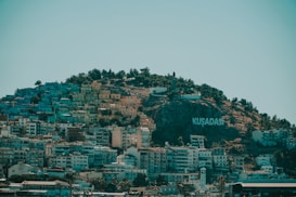 A hillside densely covered with colorful buildings with a large sign that reads 'KUŞADASI' on the right side. The top of the hill is lined with trees and a statue can be seen. The sky is clear, adding a serene backdrop to the lively urban area.