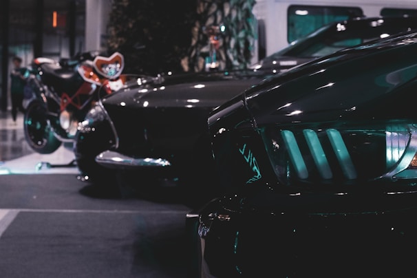 A sleek, dimly lit scene featuring a lineup of stylish vehicles, including a motorcycle and a car with prominent headlights. The setting appears to be indoors, possibly at an auto show or exhibition, with reflective surfaces and moody lighting.