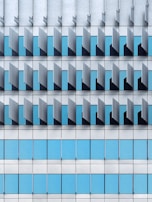 A modern architectural façade featuring a repetitive pattern of vertical and horizontal elements. The design includes blue-tinted glass windows interspersed with grey metallic panels, creating a geometric and orderly appearance.