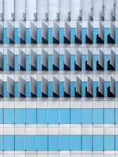 A modern architectural façade featuring a repetitive pattern of vertical and horizontal elements. The design includes blue-tinted glass windows interspersed with grey metallic panels, creating a geometric and orderly appearance.