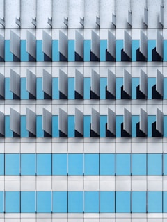 A modern architectural façade featuring a repetitive pattern of vertical and horizontal elements. The design includes blue-tinted glass windows interspersed with grey metallic panels, creating a geometric and orderly appearance.