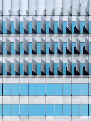 A modern architectural façade featuring a repetitive pattern of vertical and horizontal elements. The design includes blue-tinted glass windows interspersed with grey metallic panels, creating a geometric and orderly appearance.