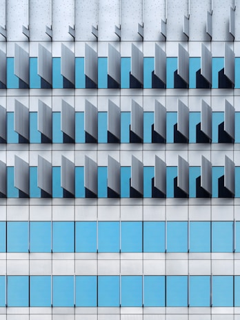 A modern architectural façade featuring a repetitive pattern of vertical and horizontal elements. The design includes blue-tinted glass windows interspersed with grey metallic panels, creating a geometric and orderly appearance.