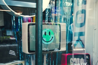 A vintage television with a green smiley face sticker displayed on the screen. The TV is situated in a shop window among various reflections and bluish curtains. Other equipment and sale signs are partially visible in the scene.