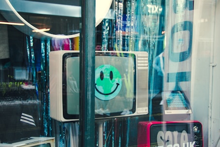 A vintage television with a green smiley face sticker displayed on the screen. The TV is situated in a shop window among various reflections and bluish curtains. Other equipment and sale signs are partially visible in the scene.