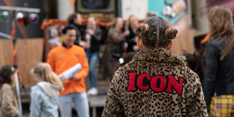 A child wearing a leopard print jacket with the word 'ICON' in bold red letters on the back is in the foreground. The scene appears to be a festive or community gathering with people socializing in the background. Decorations and some equipment are visible, suggesting an event setting.