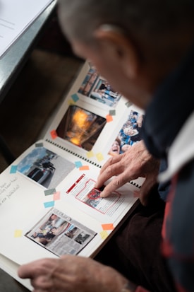 A person is looking through a scrapbook filled with various photographs and newspaper clippings. The pages have colorful paper corners holding the items in place. The images depict different scenes and articles.