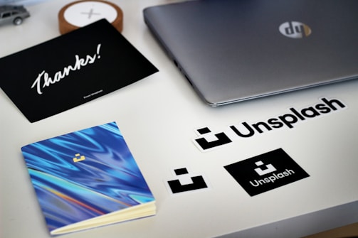 A desk with a laptop, Unsplash stickers, a notebook with a blue swirled pattern, and a card with the word 'Thanks!' written on it. The setup has a clean and modern aesthetic with a focus on branding.