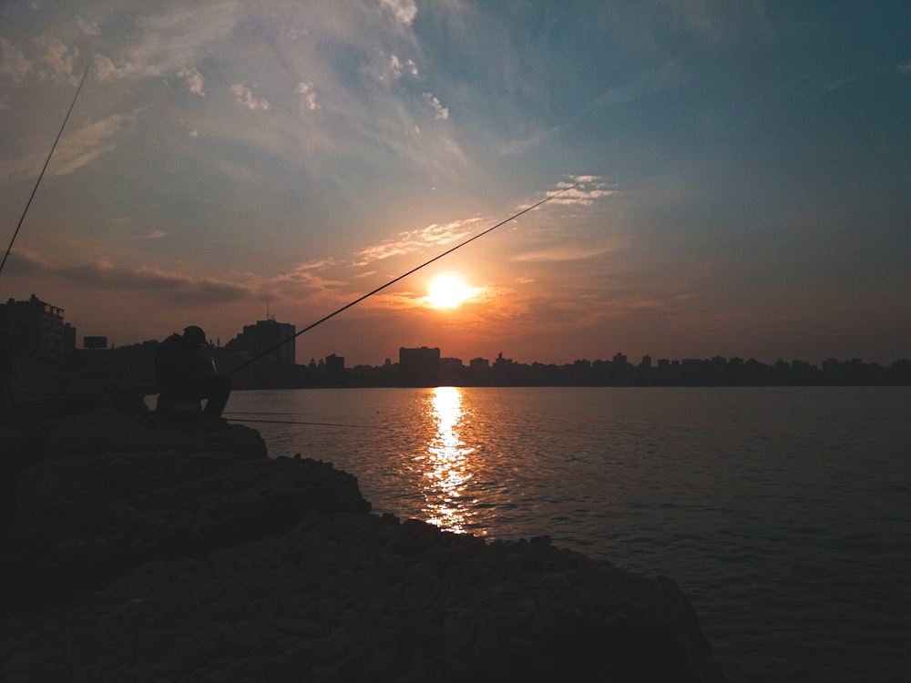 silhouette of man sitting on rock near body of water during sunset