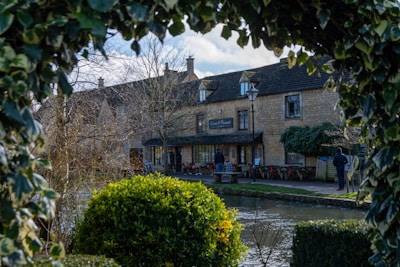 A traditional stone building with a sign indicating it is an inn or pub, surrounded by ivy and bushes. A calm stream runs in the foreground, and people are seen walking or standing nearby. The scene is framed by leafy branches, suggesting a garden setting.