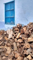 A large pile of chopped firewood is stacked against a light blue wall with a barred window. The logs are various sizes and shapes, with some pieces of paper and debris mixed in among them. The wood appears weathered and is predominantly brown in color.