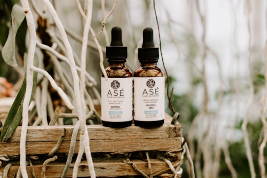 Two brown dropper bottles labeled 'As&eacute; Pure Naturals' are placed side by side on a rustic wooden surface. The bottles contain a hemp extract product and are surrounded by intertwined, dried branches, creating a natural and earthy background.