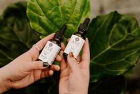 Two hands holding small bottles of Ase brand liquid, set against a backdrop of large green leaves. The bottles have black droppers and labels with text describing their contents.