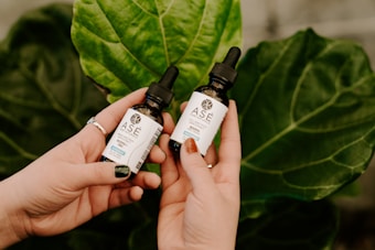 Two hands holding small bottles of Ase brand liquid, set against a backdrop of large green leaves. The bottles have black droppers and labels with text describing their contents.