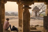 A person sits under the stone arch of a historical structure, writing in a notebook. The scene overlooks a small stone temple surrounded by barren trees against a backdrop of rocky terrain and a hazy sky.