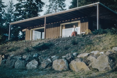 A wooden house with a sloped roof is situated on a hillside. In front of the house is a woman sitting on the porch, with large windows behind her. The hill is covered with rocks and grass, surrounded by tall trees in the background.