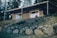 A wooden house with a sloped roof is situated on a hillside. In front of the house is a woman sitting on the porch, with large windows behind her. The hill is covered with rocks and grass, surrounded by tall trees in the background.