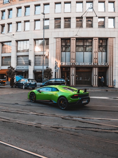 A vibrant green sports car is parked on a street in front of a building with large windows and signage that reads 'Credito Bergamasco'. The building has a modern architectural style with a series of vertical lines and is situated in an urban environment with tram tracks visible on the road. A few other cars are parked nearby, and pedestrians can be seen in the background.
