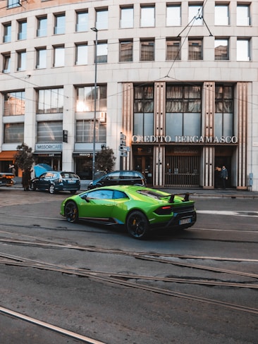 A vibrant green sports car is parked on a street in front of a building with large windows and signage that reads 'Credito Bergamasco'. The building has a modern architectural style with a series of vertical lines and is situated in an urban environment with tram tracks visible on the road. A few other cars are parked nearby, and pedestrians can be seen in the background.