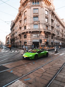 A vibrant green sports car is driving down a busy city street with an ornate, historic building in the background. The street features tram tracks, and there are people on bicycles, motorcycles, and a pedestrian crossing the road. The area has a mix of modern transportation amidst classic architecture.