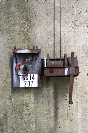 Two metal objects mounted on a concrete wall, including a rusty mechanical device with a handle and a rectangular metal panel with pipes and a number plate reading 'SC14 207'.