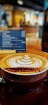 A cup of coffee with intricate latte art on top, placed on a wooden table in a café setting. In the background, there is a blurred menu board and the warm, cozy interior supports the relaxed ambiance.