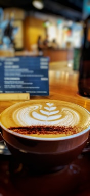 A cup of coffee with intricate latte art on top, placed on a wooden table in a café setting. In the background, there is a blurred menu board and the warm, cozy interior supports the relaxed ambiance.