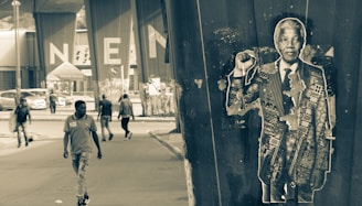 grayscale photo of man in black and white camouflage jacket standing beside wall