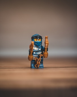 A small LEGO ninja figurine stands on a wooden surface, dressed in a dark blue outfit with matching headgear. It holds nunchucks in its hands, and its facial expression appears determined.