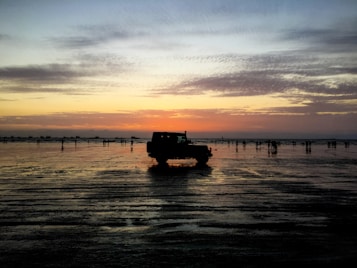 A silhouette of a jeep is visible on a wet beach during sunset. The sky is a blend of orange, yellow, and blue shades, with scattered clouds. The reflection of the jeep and people walking is noticeable on the wet sand, creating a serene and picturesque scene.