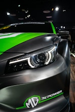 A close-up view of the front section of a sleek car, featuring a prominent headlight design with LED elements. The car has a black and green color scheme with the logo 'MG X POWER' at the front. The car is positioned on a wooden floor, and the lighting suggests it is displayed at an indoor event or showroom.