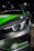 A close-up view of the front section of a sleek car, featuring a prominent headlight design with LED elements. The car has a black and green color scheme with the logo 'MG X POWER' at the front. The car is positioned on a wooden floor, and the lighting suggests it is displayed at an indoor event or showroom.