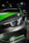 A close-up view of the front section of a sleek car, featuring a prominent headlight design with LED elements. The car has a black and green color scheme with the logo 'MG X POWER' at the front. The car is positioned on a wooden floor, and the lighting suggests it is displayed at an indoor event or showroom.