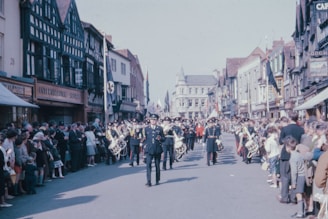 A marching band is parading down a wide street flanked by rows of spectators. The band members are in uniform, playing instruments such as drums and brass. The street is lined with historic buildings featuring half-timbered facades. People of various ages are gathered, watching the procession under a clear sky.