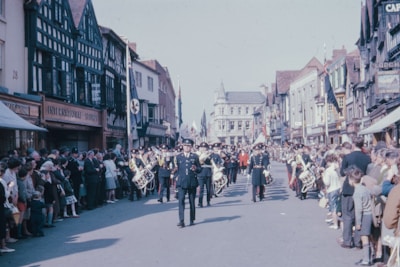 A marching band is parading down a wide street flanked by rows of spectators. The band members are in uniform, playing instruments such as drums and brass. The street is lined with historic buildings featuring half-timbered facades. People of various ages are gathered, watching the procession under a clear sky.